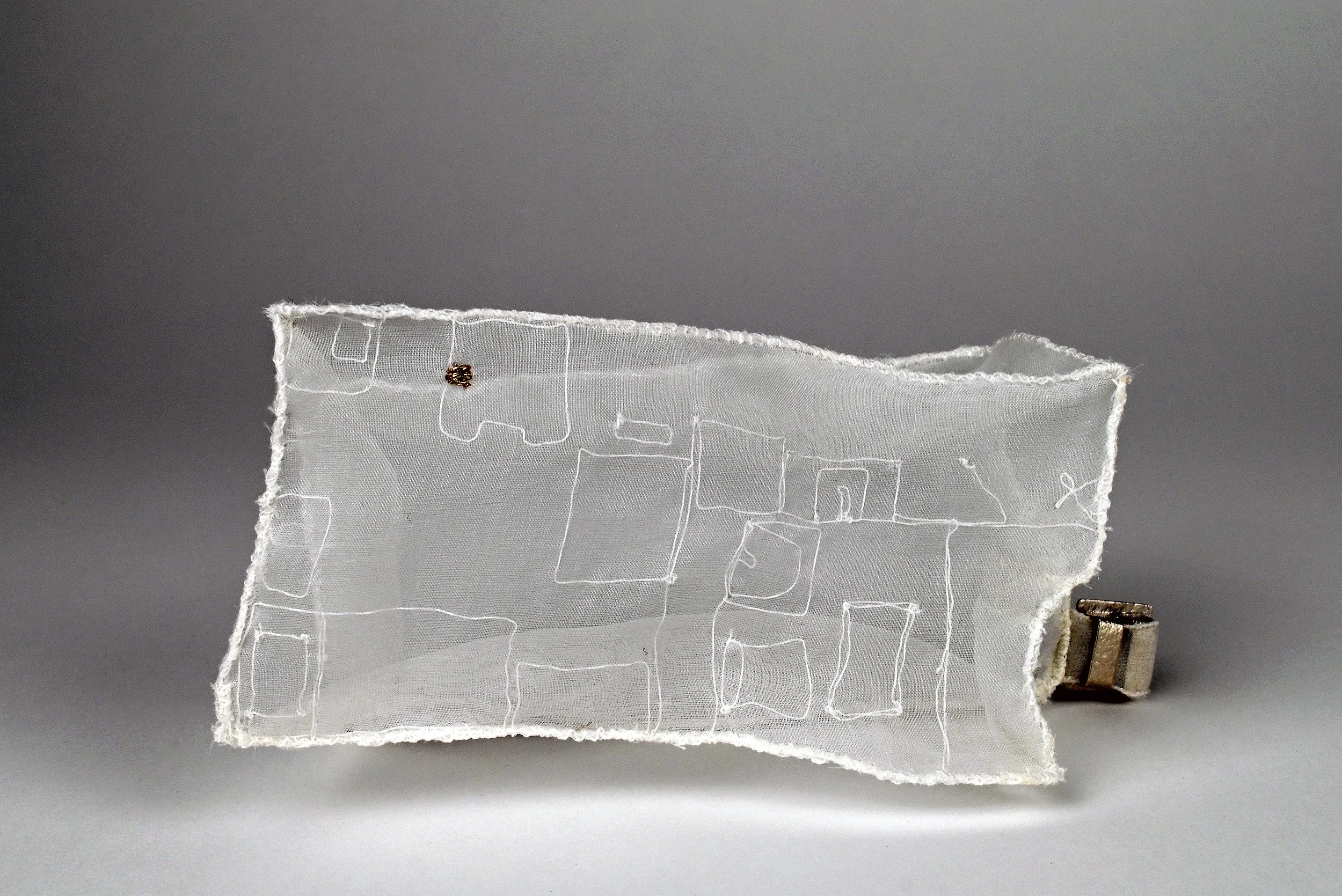 A piece of white fabric with stitched outlines of windows and doors, attached to a metal clip, with a small goldnen mark on it, against a plain gray background.
