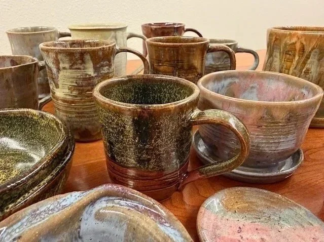 Collection of handmade ceramic mugs and bowls with earthy glaze finishes arranged on a wooden surface.