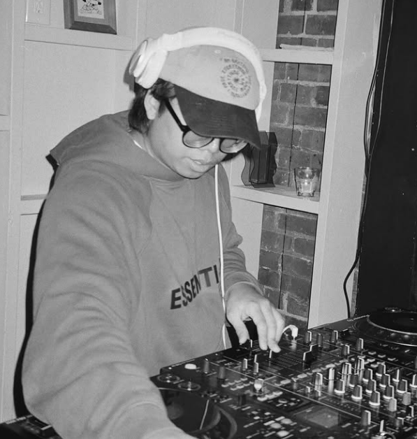 A person wearing a baseball cap, glasses, headphones, and a hoodie is DJing with a mixer and turntables in a cozy indoor setting.