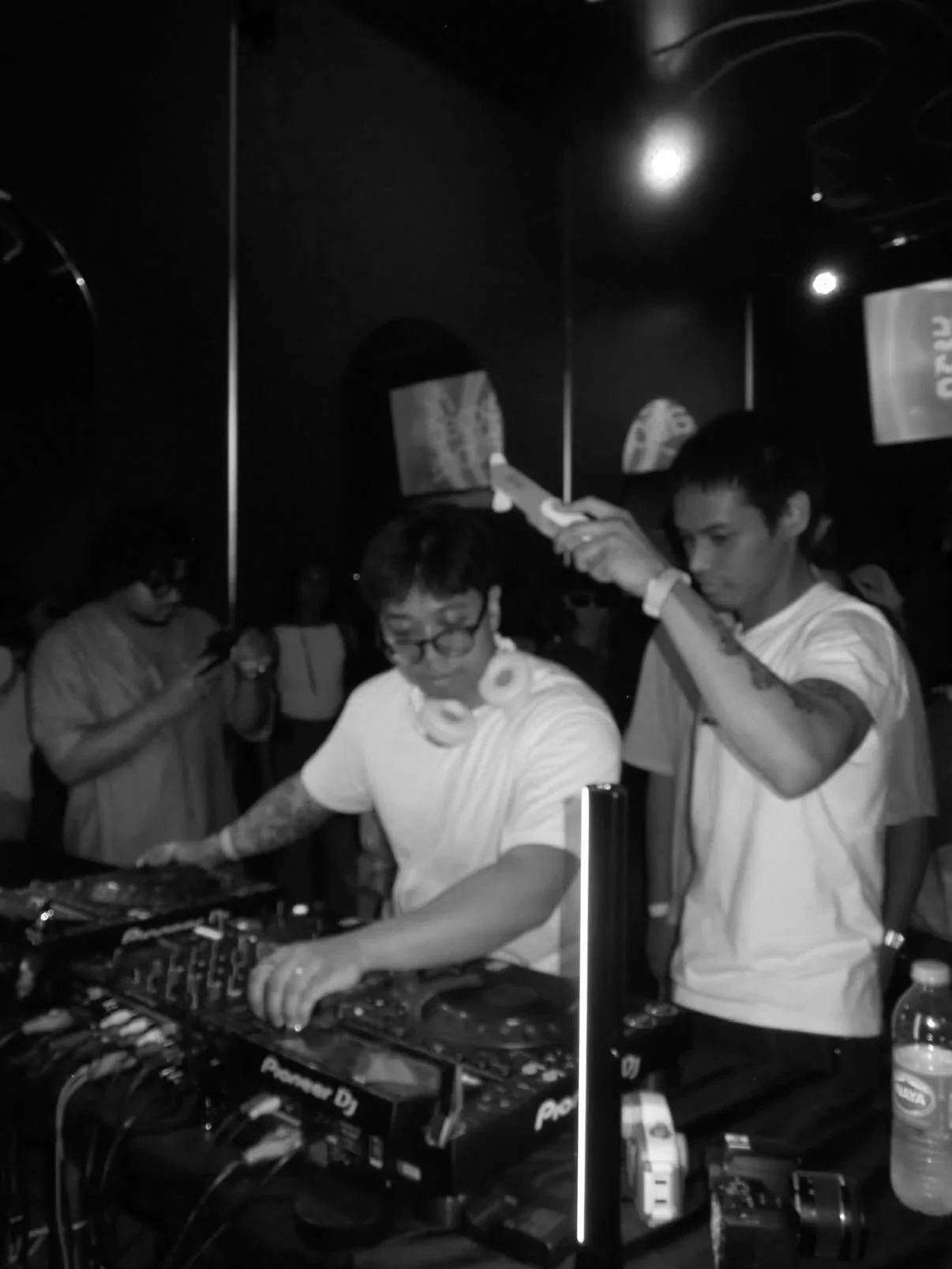 Two young men are DJing at a club, one is operating a turntable and the other is adjusting equipment. The crowd is watching and one person is taking a photo in the background.
