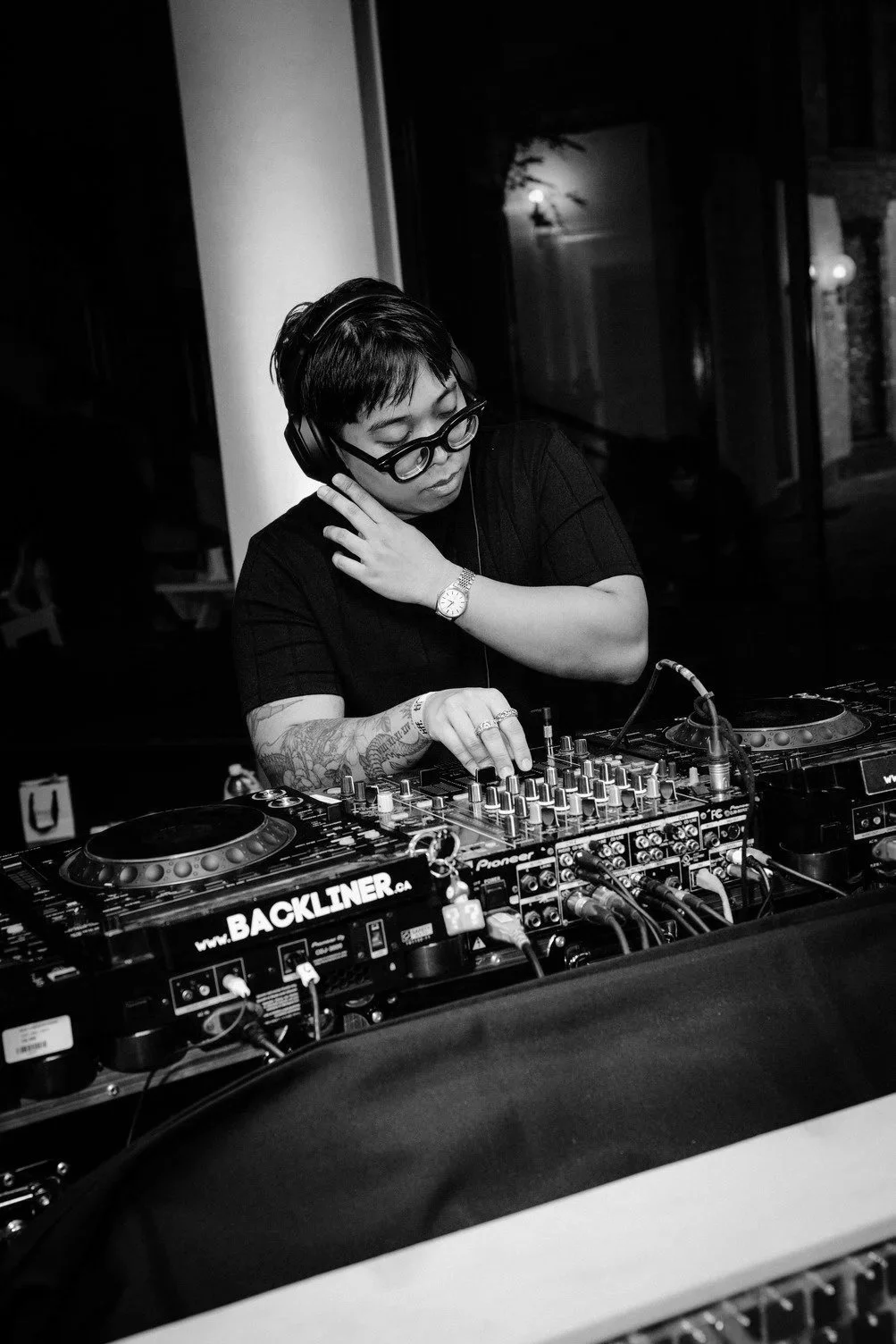 A woman with short hair, glasses, and tattoos adjusts music controls on a DJ mixer at night in black and white.