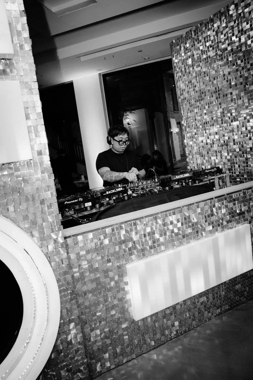 A DJ, wearing glasses and headphones, standing behind a DJ booth with equipment, in a room with tiled walls.