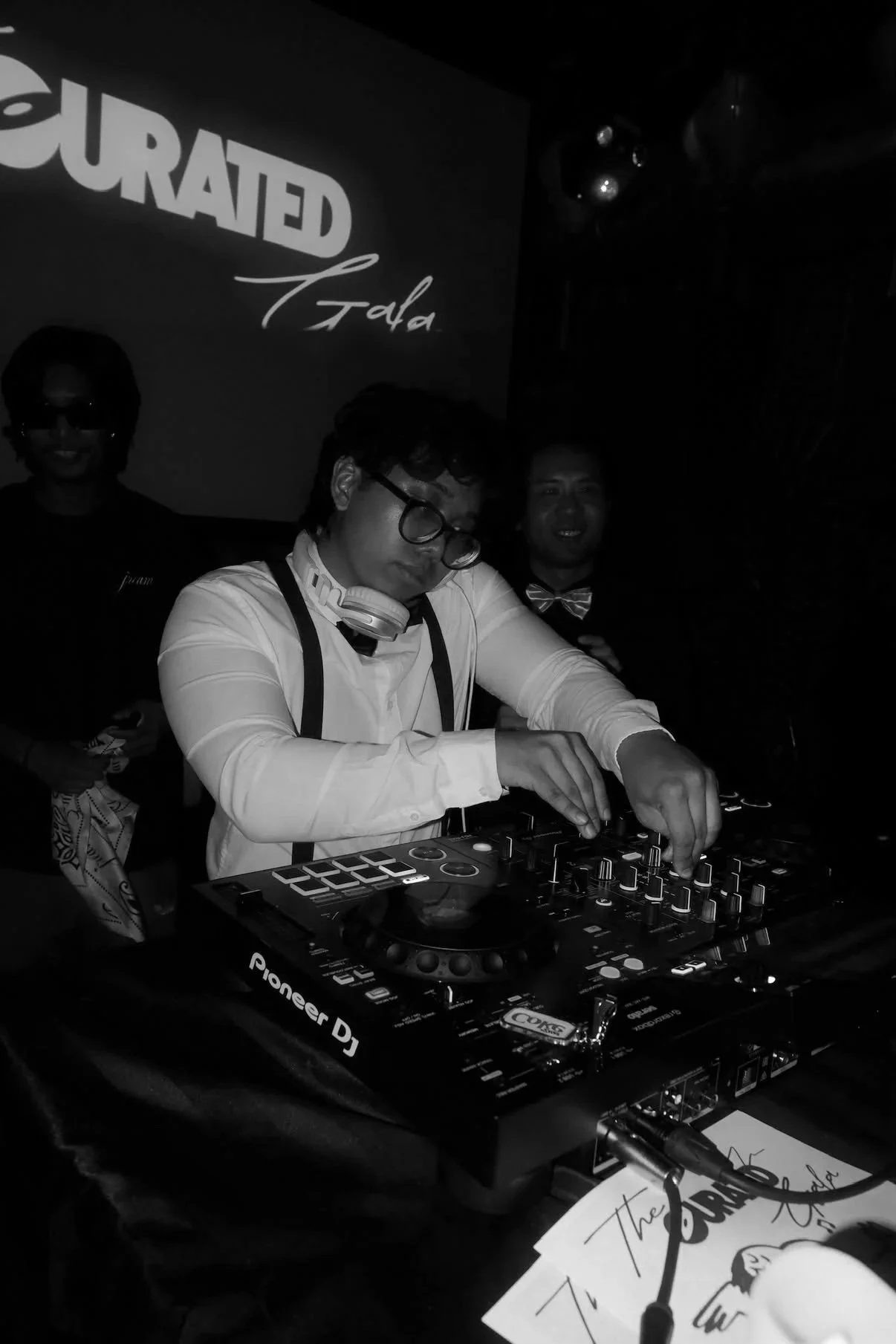 A DJ wearing glasses, a white shirt, and headphones around his neck DJing at a party, with two men in the background, one dressed in a black shirt and the other in a tuxedo with a bow tie, in a dark setting with a large screen displaying the word 'CURATED' and 'Fida' behind them.
