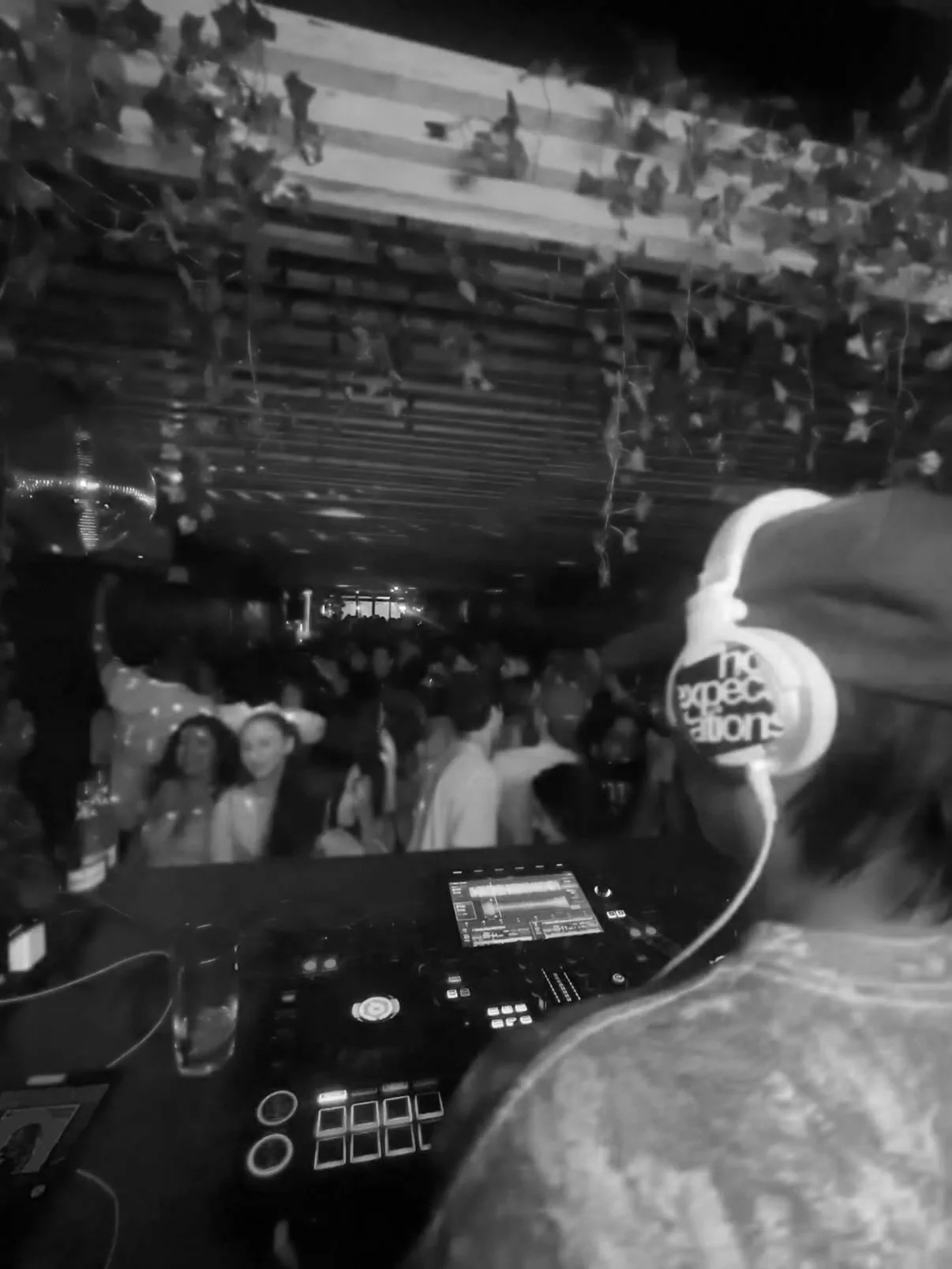 A DJ performing at an event with a crowd of people dancing and socializing in a dimly lit indoor venue. The DJ's equipment is visible in the foreground, and there are decorations hanging from the ceiling.