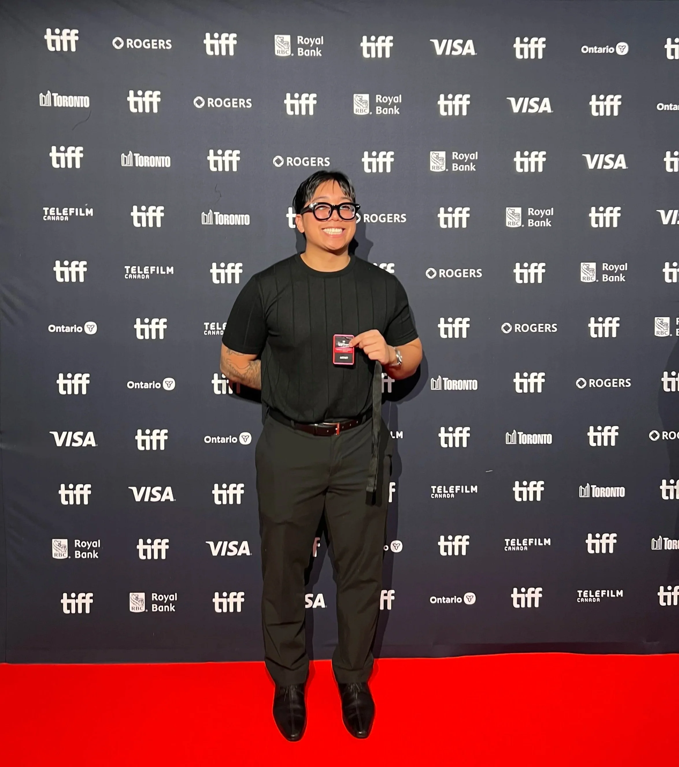 A person with short dark hair, glasses, and a big smile, standing in front of a step-and-repeat backdrop at a film festival, wearing a black shirt, black pants, and black shoes, holding a red ticket or card in their right hand, and standing on a red carpet.