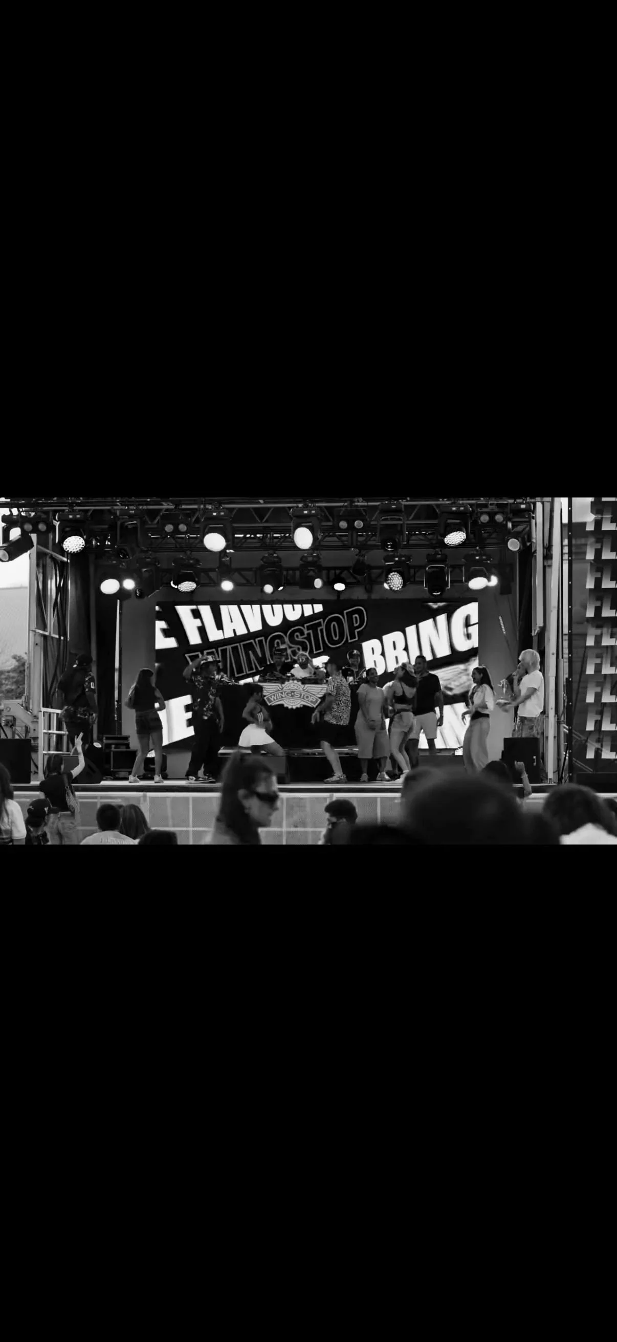 People dancing on an outdoor stage with a large screen behind them displaying the words "Eat Flavor, Wing Stop, Bring" in bold letters, with stage lighting overhead.