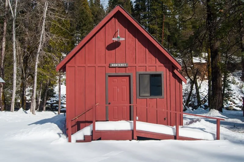 A red wooden cabin with a ramp, surrounded by snow and trees, with a sign labeled 'Monterey' above the door.