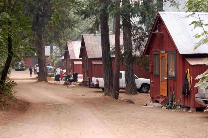 Row of red cabins with metal roofs along a dirt road in a wooded area, with people and vehicles parked nearby.