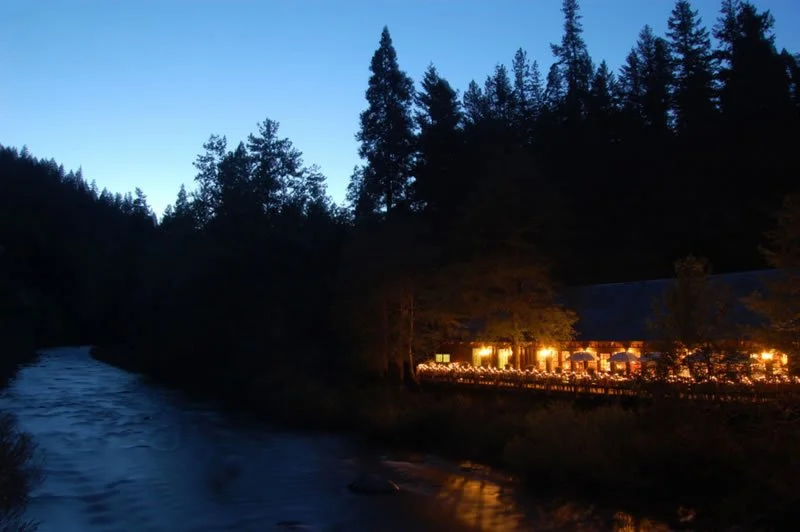 A nighttime scene of a river with a wooded area and tall pine trees. On the right, there is a building with warm lights, possibly a restaurant or lodge, nestled among trees.