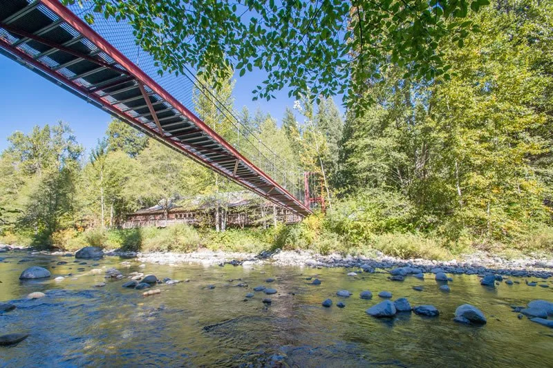 Red suspension bridge over a rocky river in a forested area with trees and a building in the background.