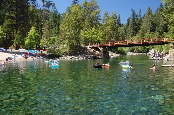 People swimming and floating on inflatable tubes in a clear green river, with a bridge and trees in the background on a sunny day.
