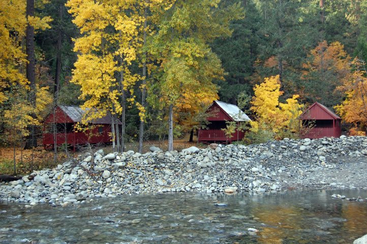Three red wooden cabins on a rocky shoreline with fall-colored trees in the background.