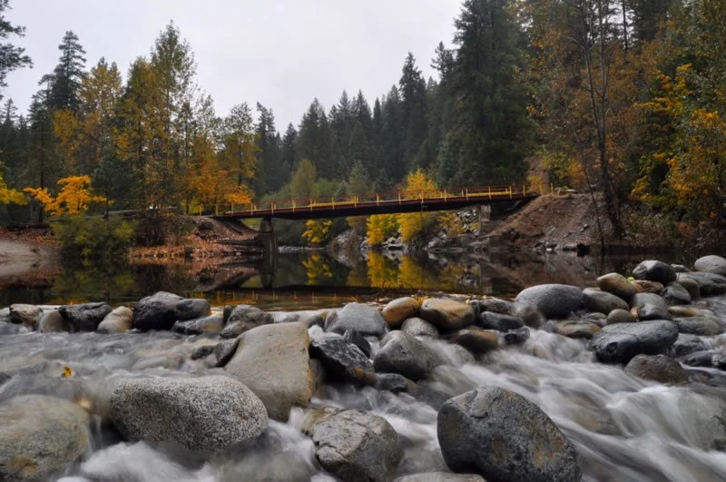 A river flowing over rocks with a bridge and trees in autumn colors in the background.