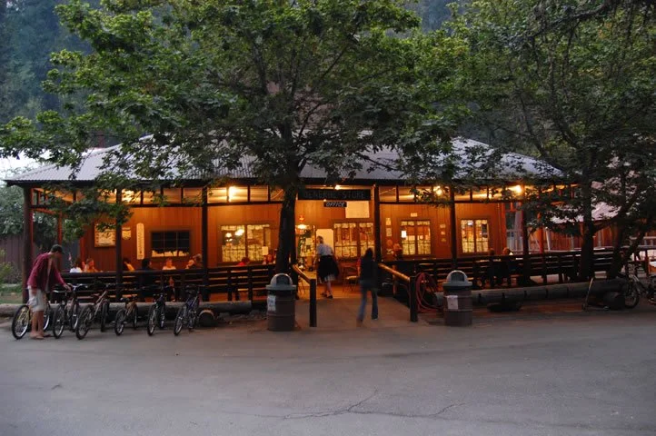 People walking and biking in front of a warmly lit, wooden rustic building with trees in the foreground.
