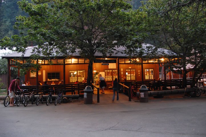 A cozy outdoor dining area with a wooden building, surrounded by trees, with people sitting and walking outside in the evening.