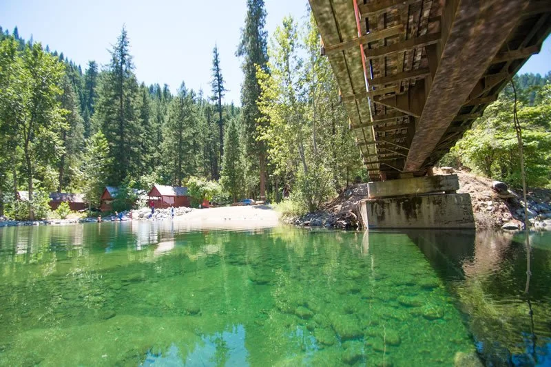 A clear green lake under a wooden bridge in a forest with trees and small cabins in the background.