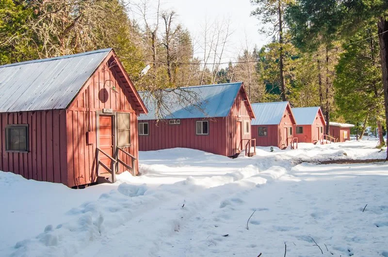 Four red wooden cabins with gray roofs in a snowy forested area.