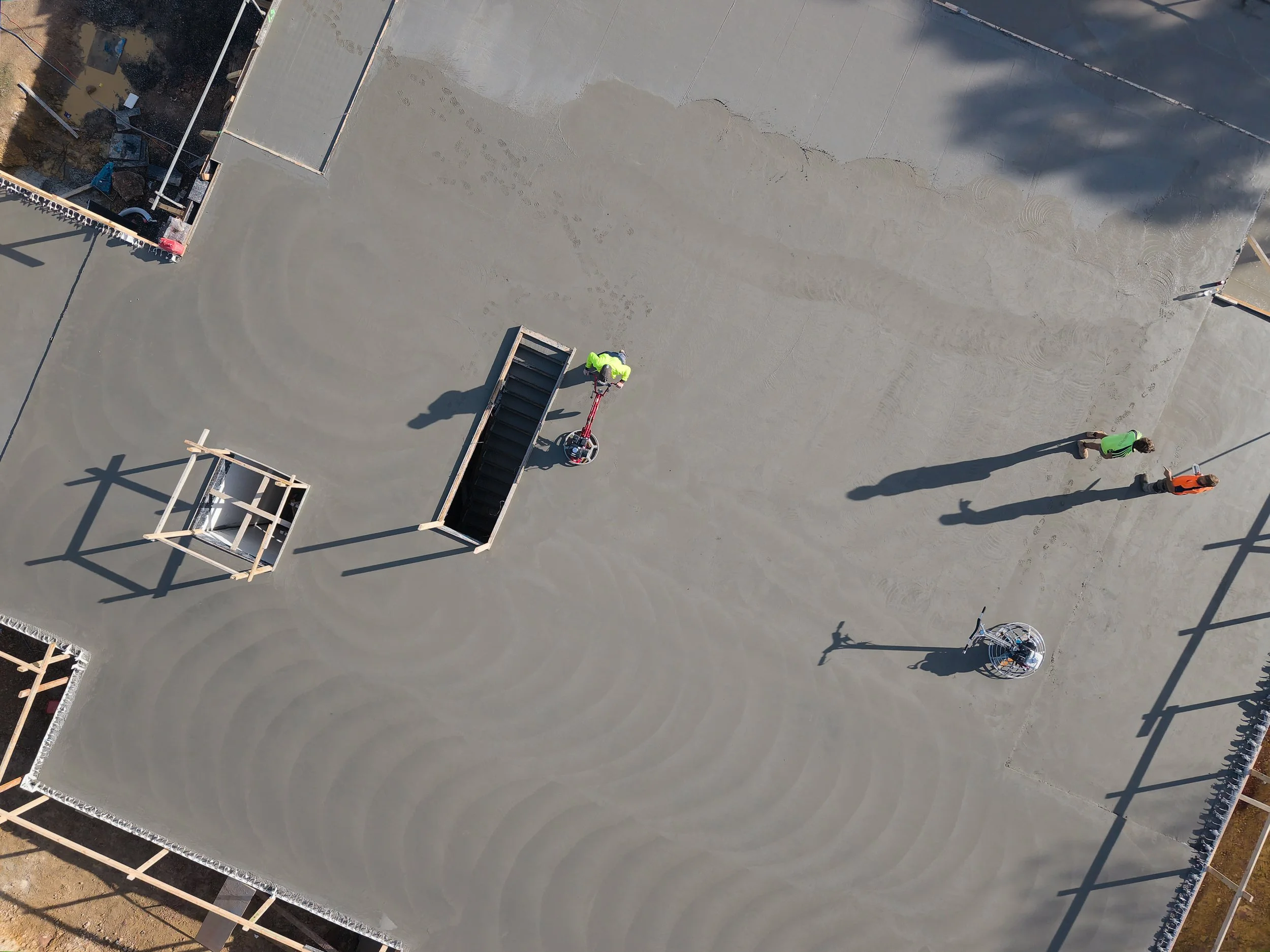 An aerial view of a construction site with workers laying concrete on a flat surface, surrounded by building frames and construction materials. Shadows cast by structures and workers are visible.