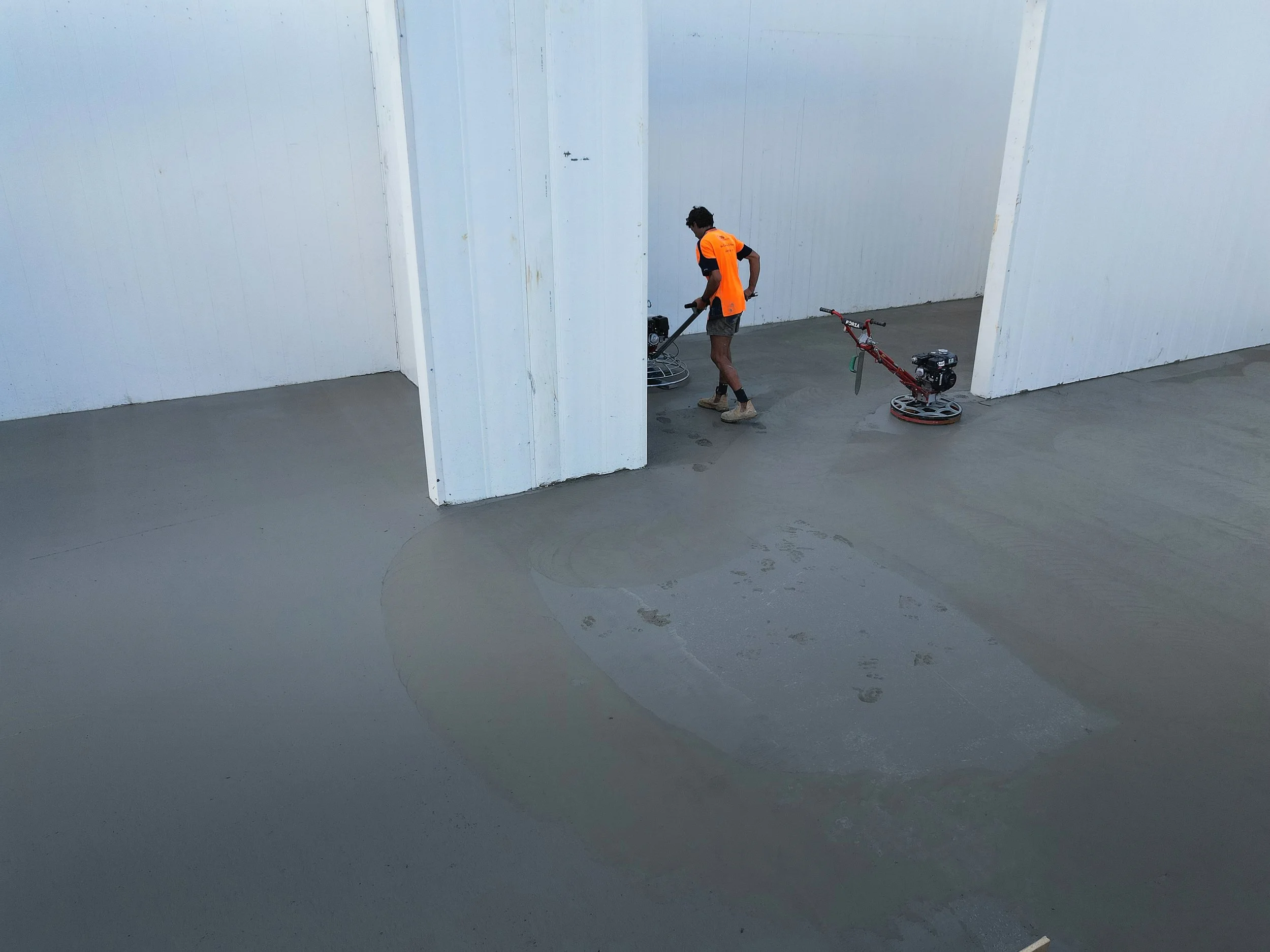 Worker in an orange vest operating a power trowel on a concrete floor, with another power trowel nearby ion a freshly poured concrete slab.