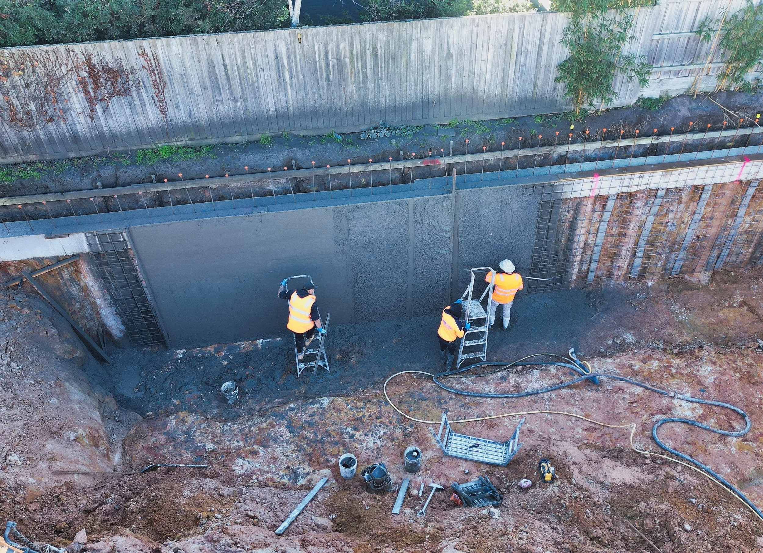Construction workers in orange vests working on a concrete basement in an excavated site, with rebar and tools visible. A wooden fence and some greenery are in the background.