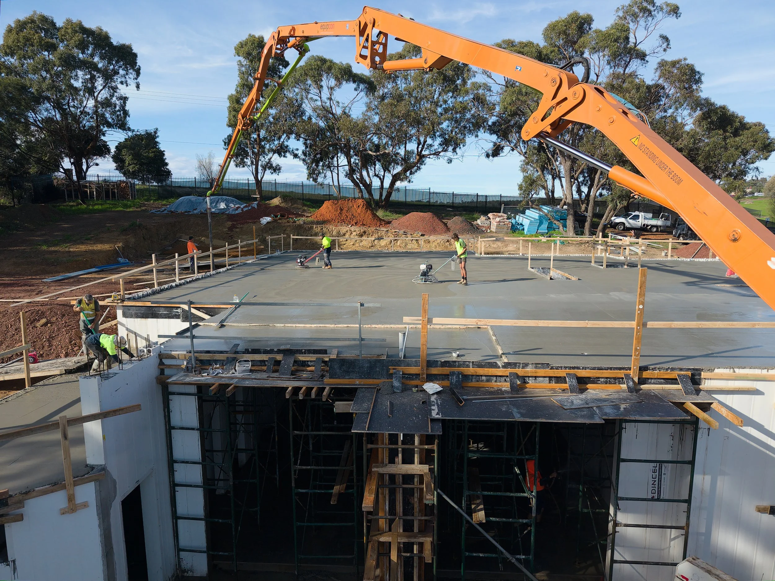Construction workers smoothing wet concrete on a building foundation with a large orange concrete pump extending overhead. The construction site is outdoors with trees and a fence in the background.