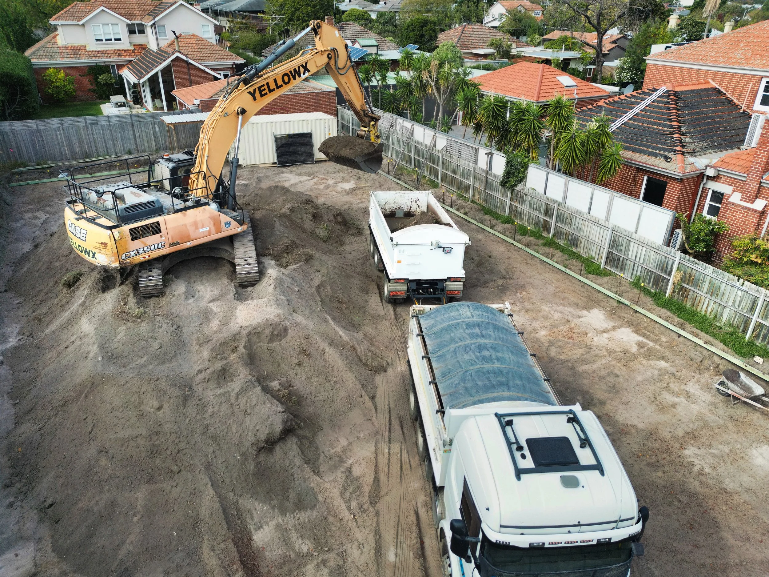 Construction site shows a yellow excavator removing dirt into a white dump truck, with neighboring houses and trees in the background.