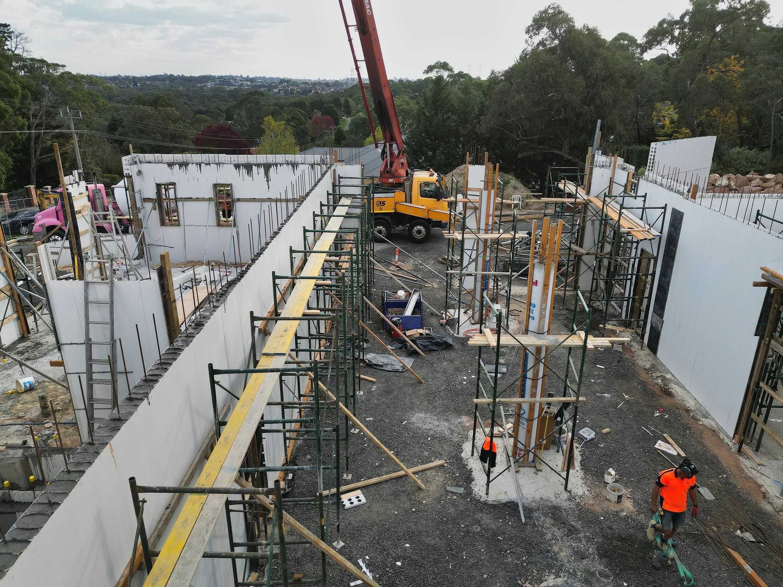 Construction site with partially built dencil concrete walls, scaffolding, a crane, and workers wearing orange vests, set against a backdrop of trees and distant cityscape.