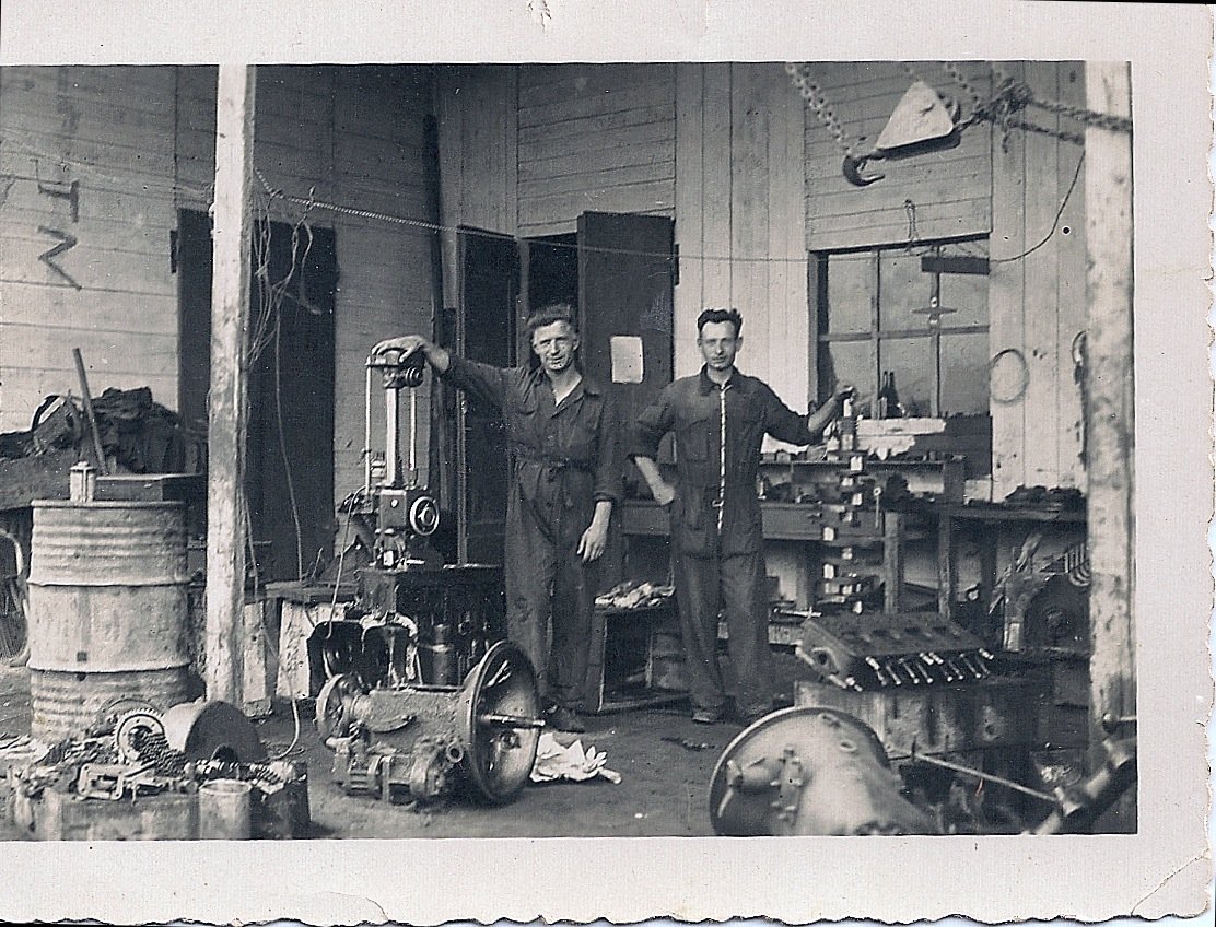 Two men in work overalls standing in a workshop surrounded by mechanical parts and tools.