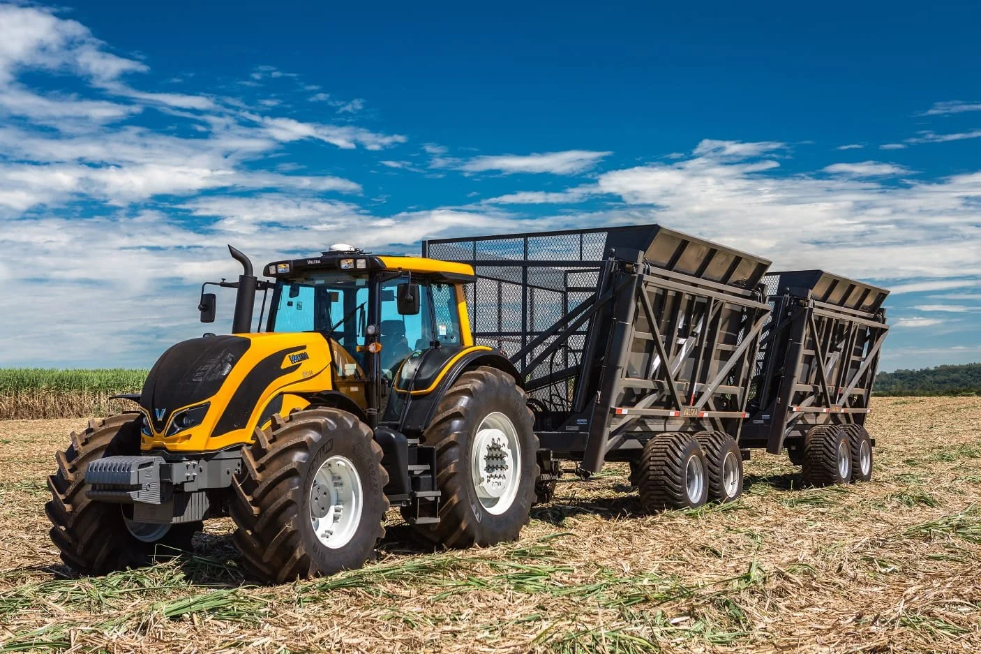 A yellow and black Valtra with attached wagons in a field under a blue sky with clouds.