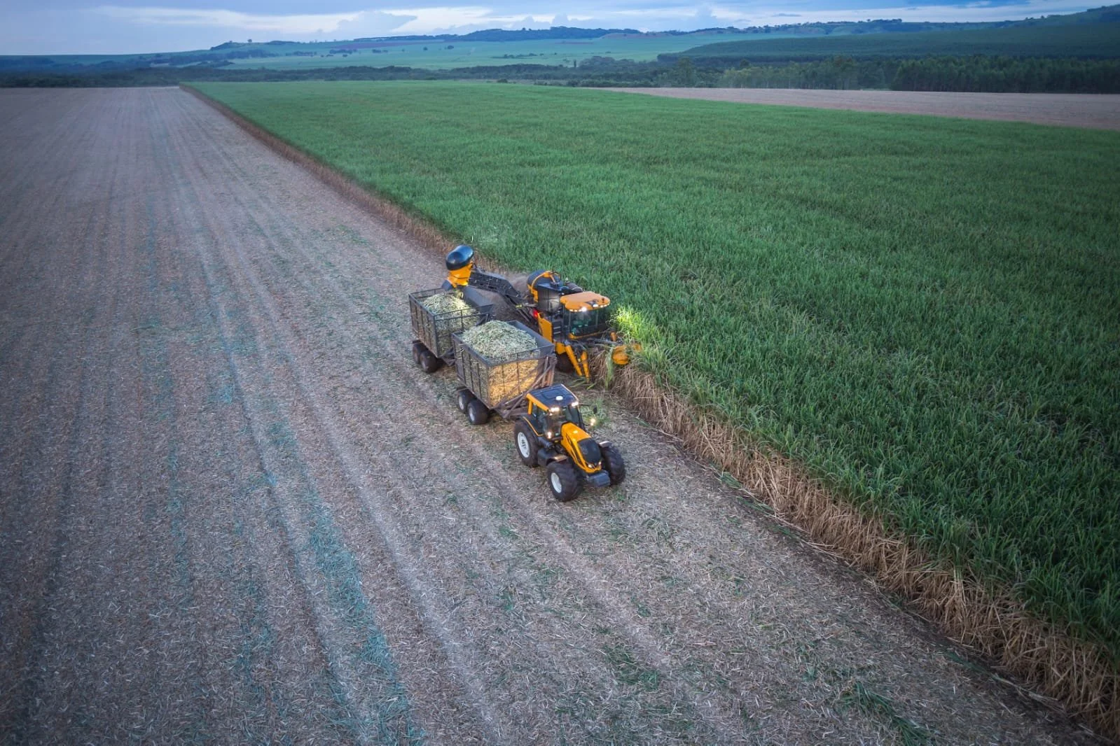 An aerial view of a Valtra harvesting crops in a large field with green crops on one side and a harvested area on the other, under a cloudy sky.