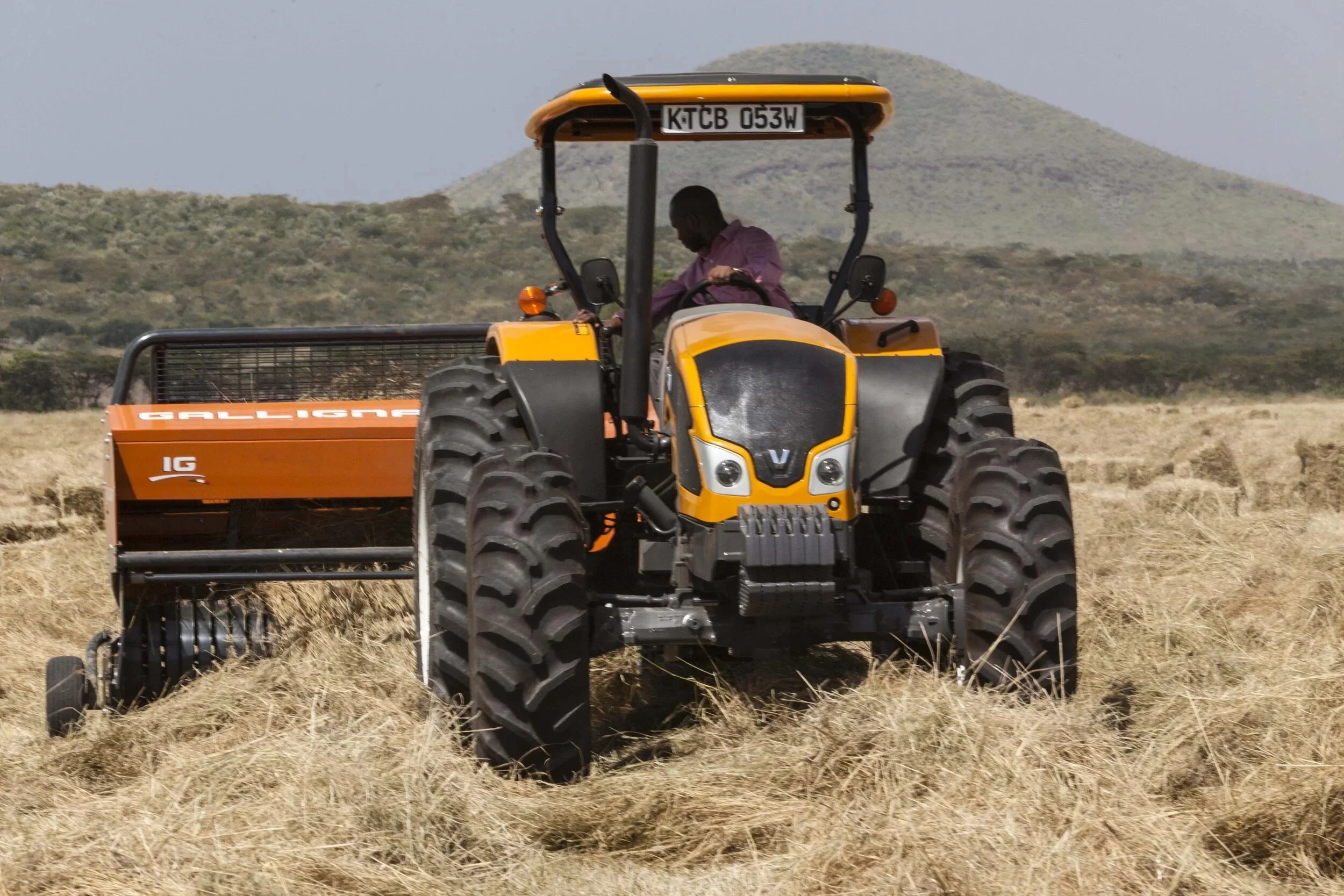 A man wearing a purple shirt is driving a yellow Valtra across a dry field with mountain in the background.