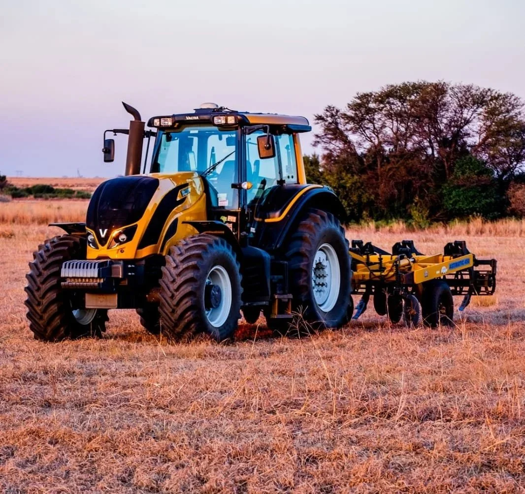 A Valtra working on a dry, grassy field with trees in the background.