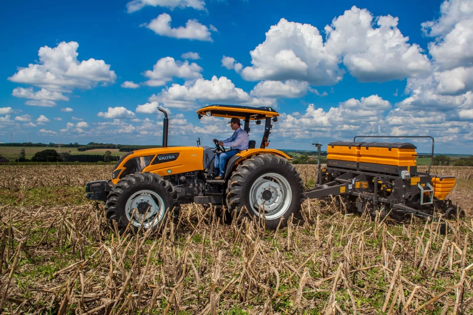 Farmer driving a yellow Valtra in a field under a cloudy blue sky, with harvested crops and farm landscape in the background.