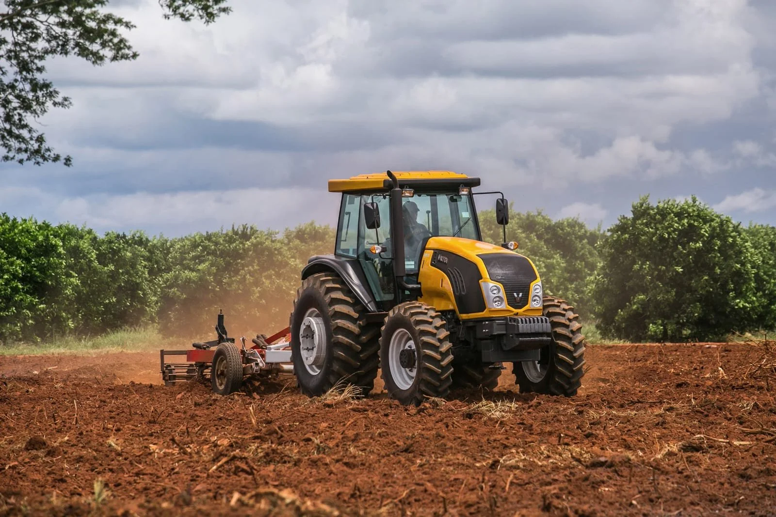 A yellow valtra working on a farm field under cloudy sky, with green trees in the background.