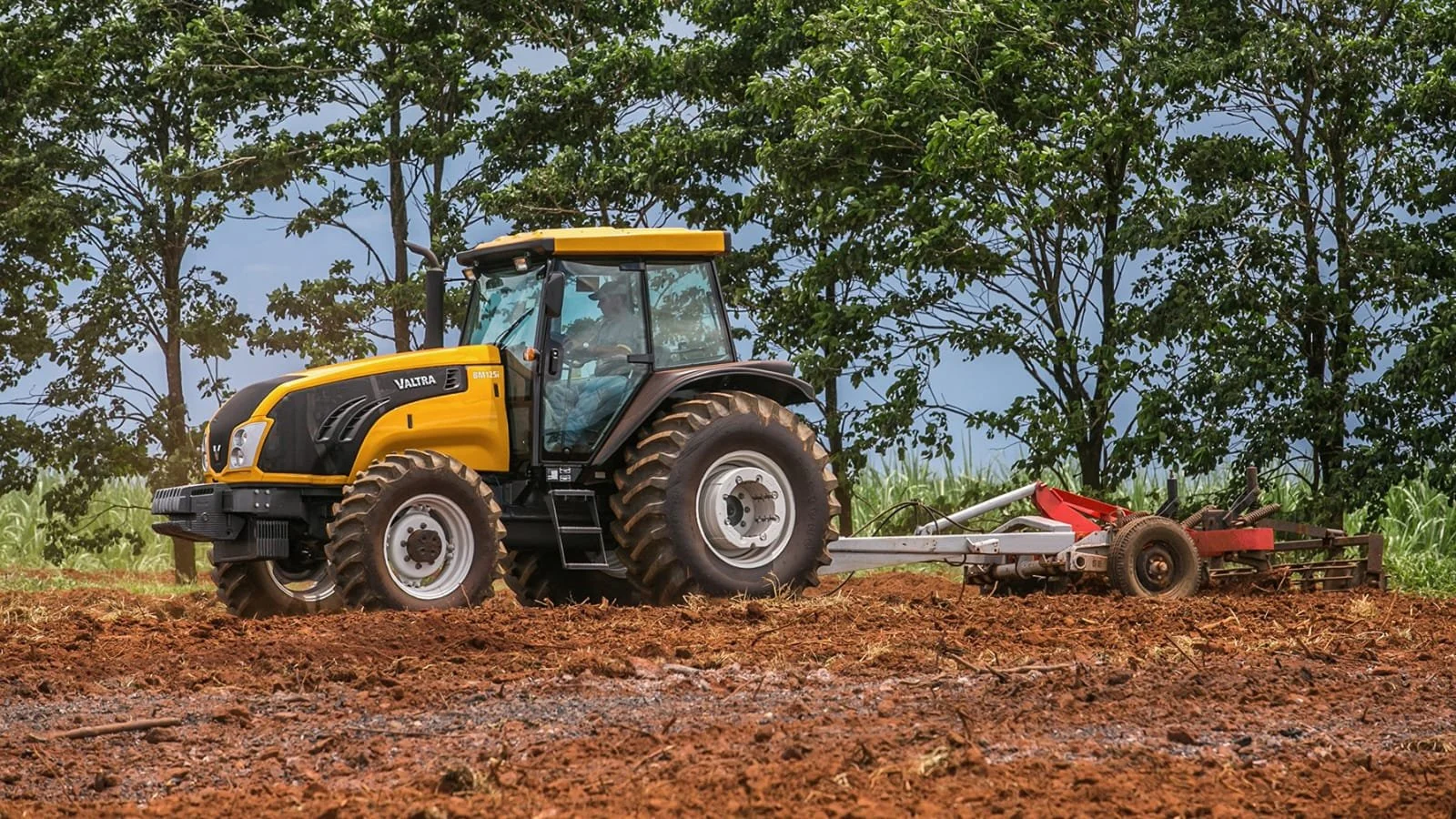 A yellow and black Valtra plowing a field with trees and a cloudy sky in the background.