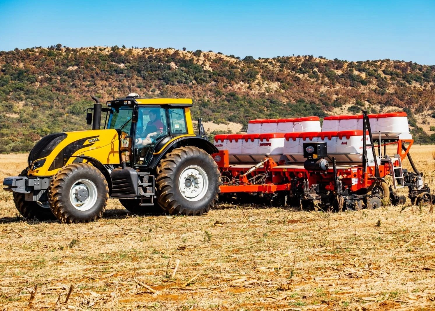 Yellow and black Valtra pulling a red and white agricultural implement in a dry field with hills in the background.