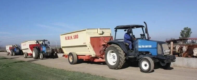 A Valtra pulling a trailer with a worker seated inside, hauling a large container on a rural road with more Valtras and trees in the background.