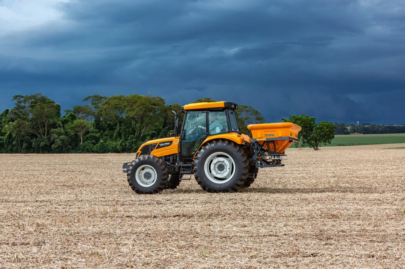 A yellow Valtra in a plowed field with dark storm clouds overhead and trees in the background.