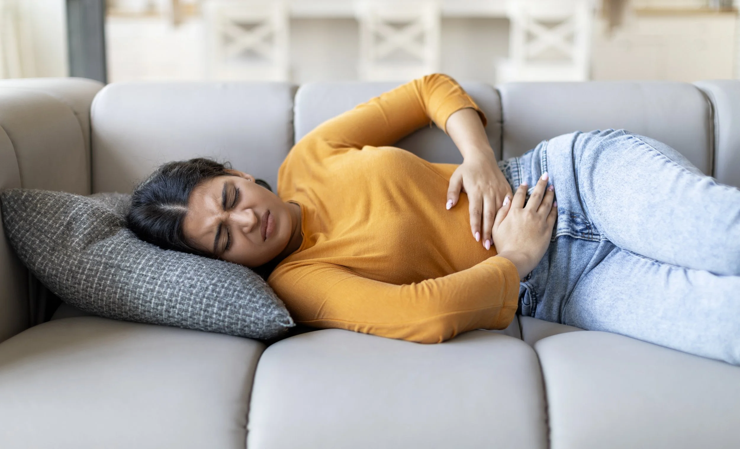 A woman lying on a couch, holding her stomach, appearing to experience discomfort, in a brightly lit room.