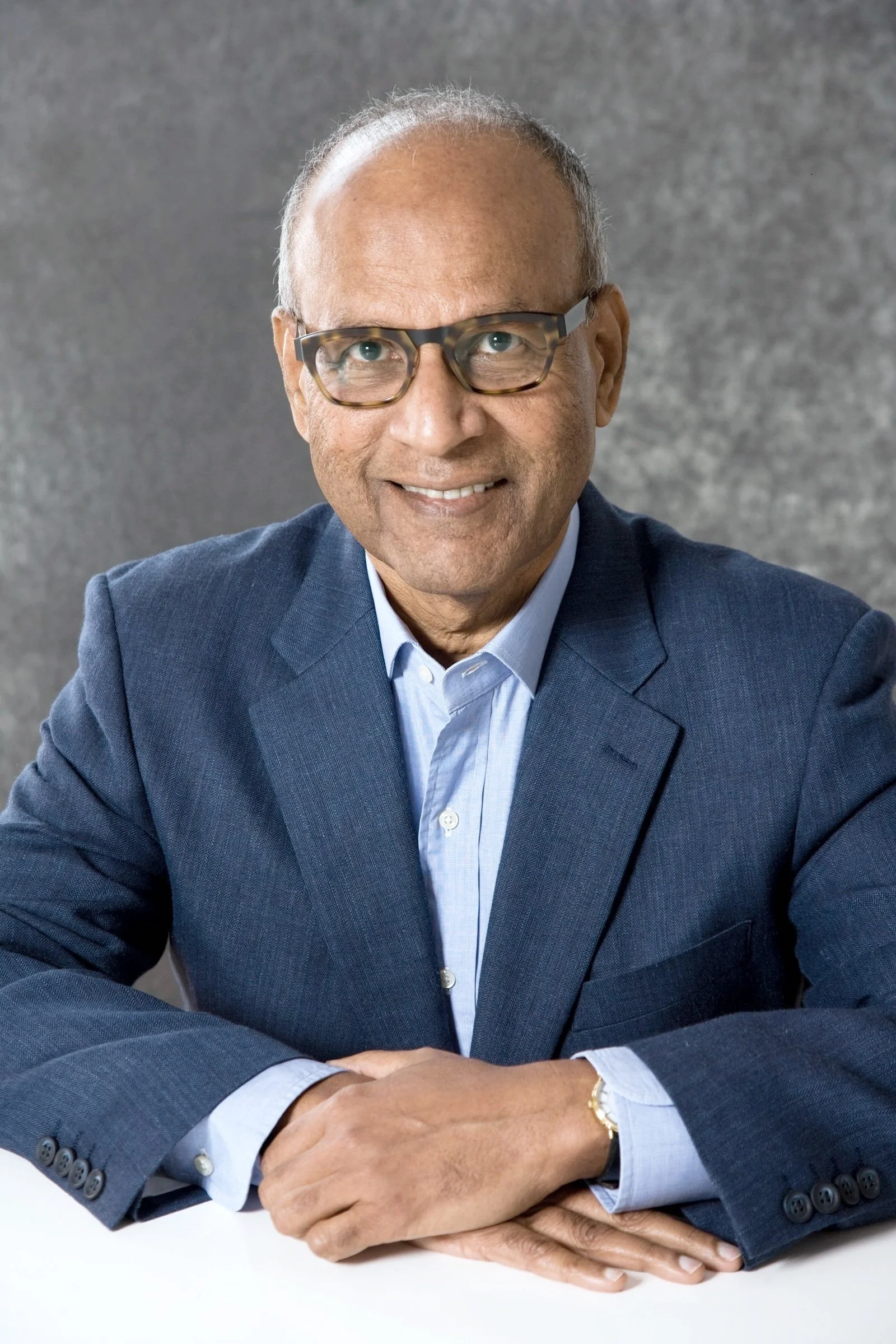Dr Iggy: A mature man with glasses, dressed in a blue suit and light blue shirt, sitting with his arms crossed on a white surface, smiling against a gray textured background.