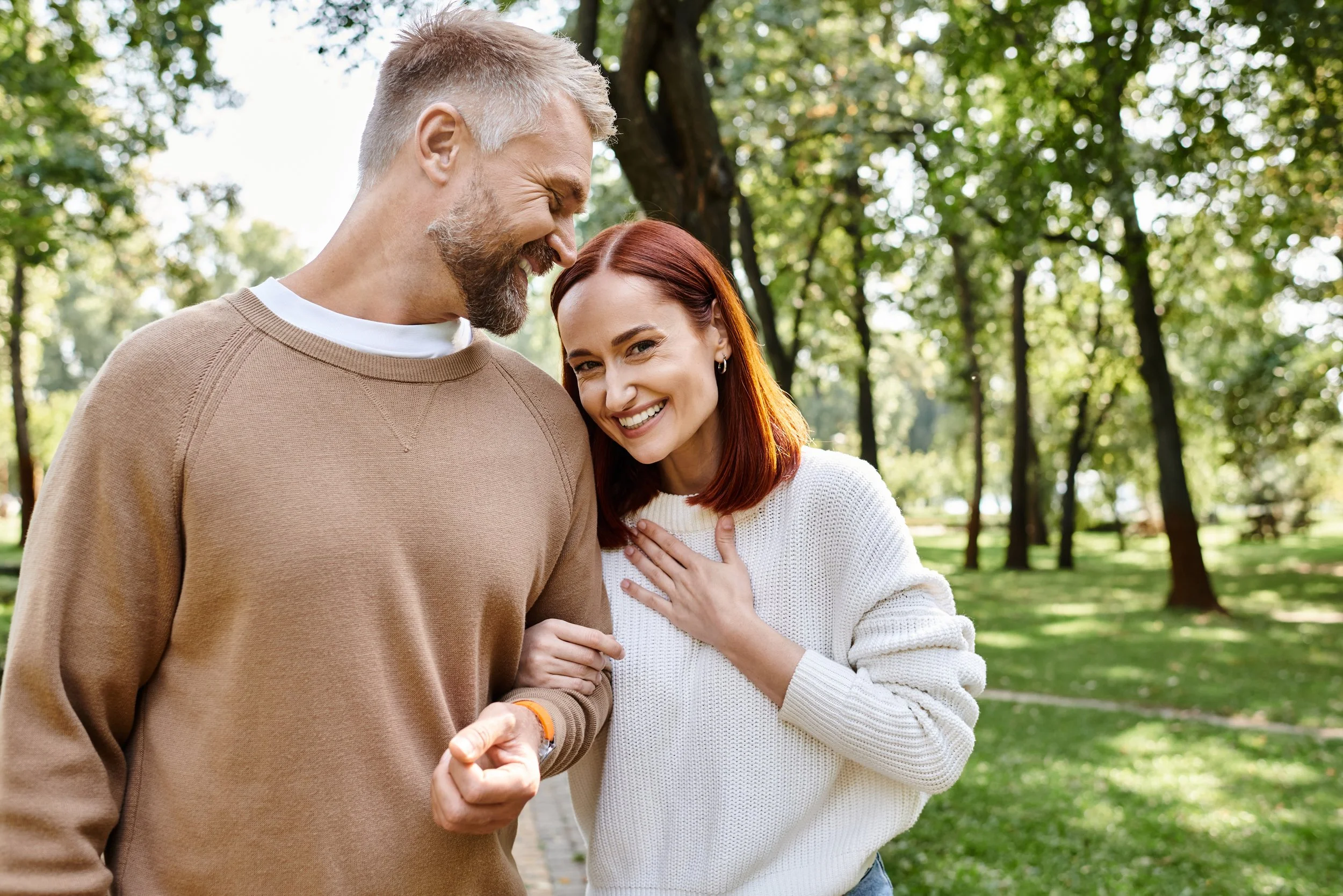 A man and woman smiling and holding hands outdoors in a park with trees in the background.