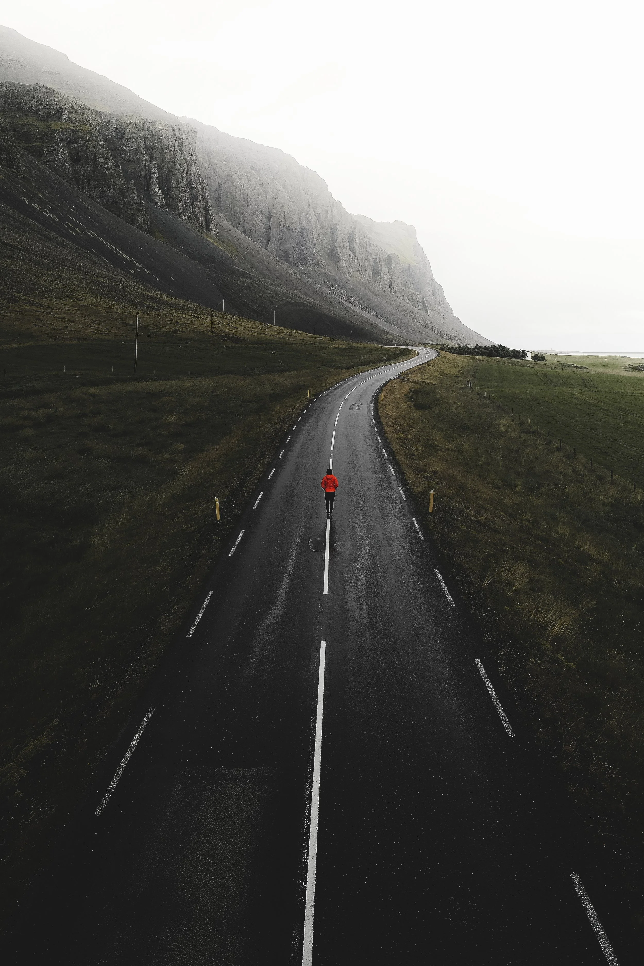 A person in a red jacket walking on a winding, empty mountain road with steep cliffs on one side and a green field on the other, under a cloudy sky.
