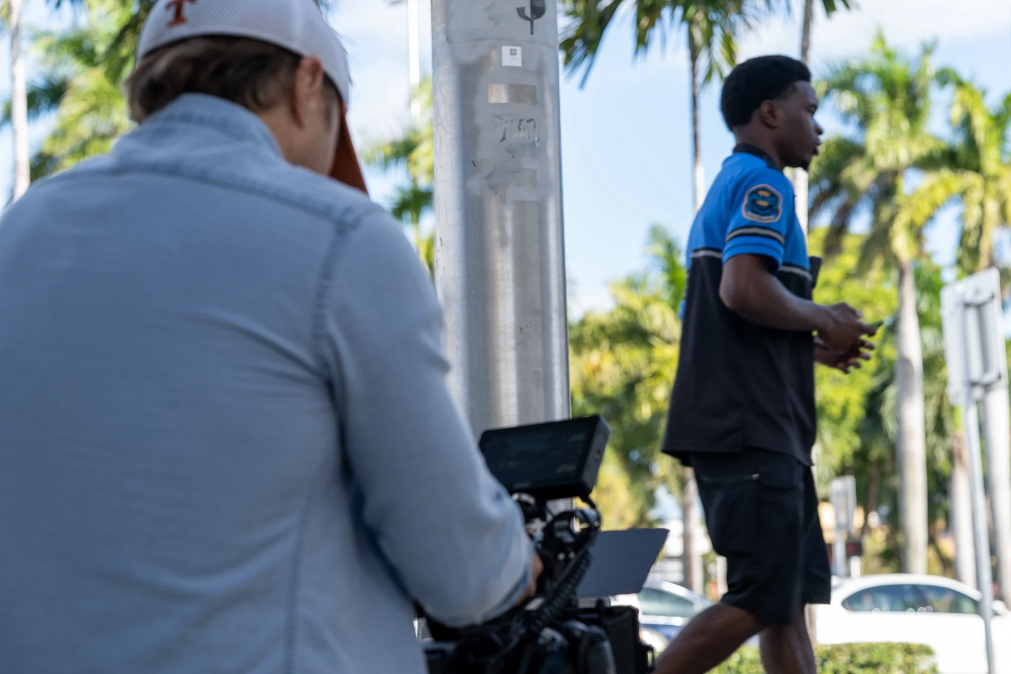 A person operating a camera captures a man walking on a sidewalk with palm trees in the background.