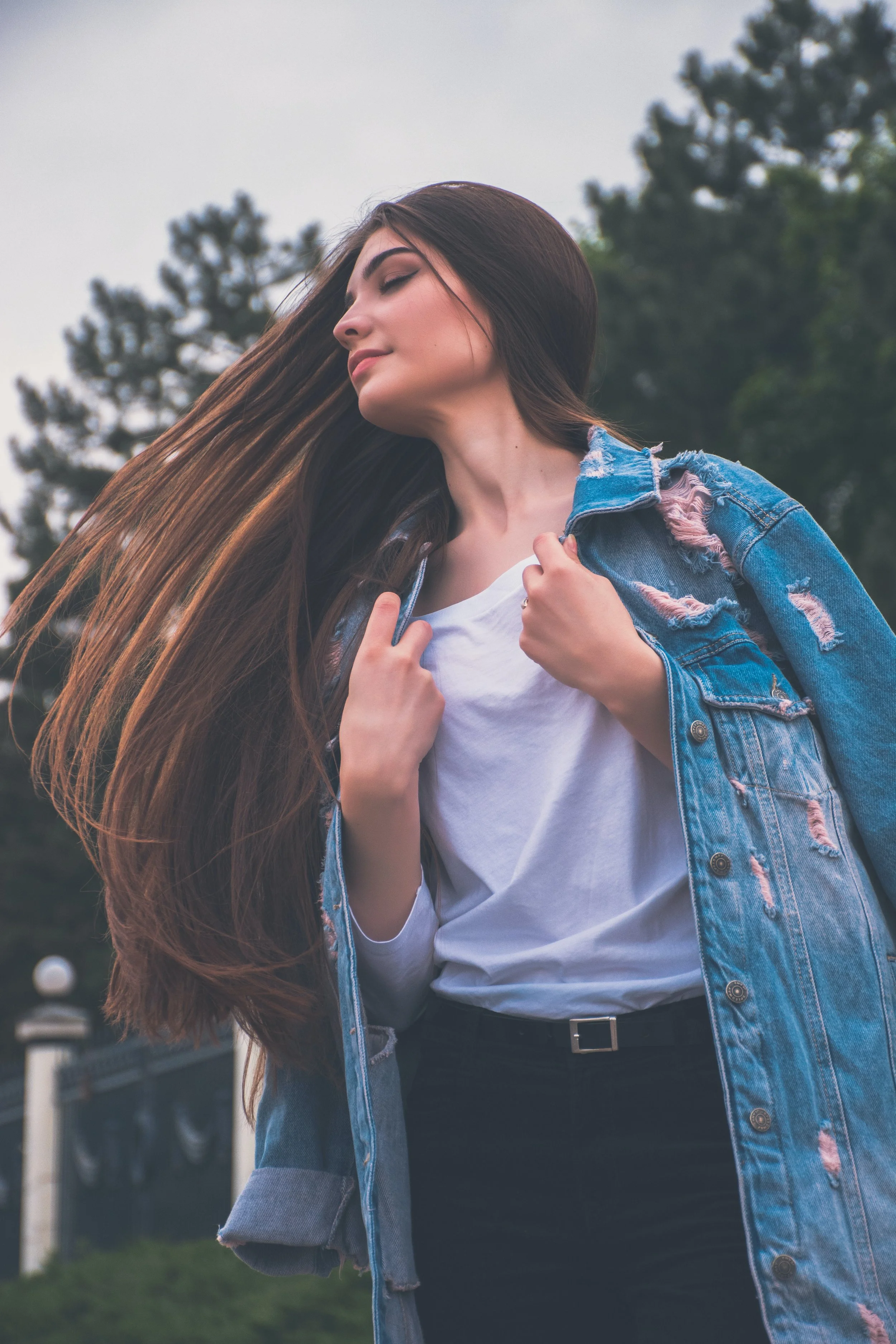 Young woman outdoors with long brown hair, wearing a distressed denim jacket over a white t-shirt, standing on a grassy area with trees in the background.
