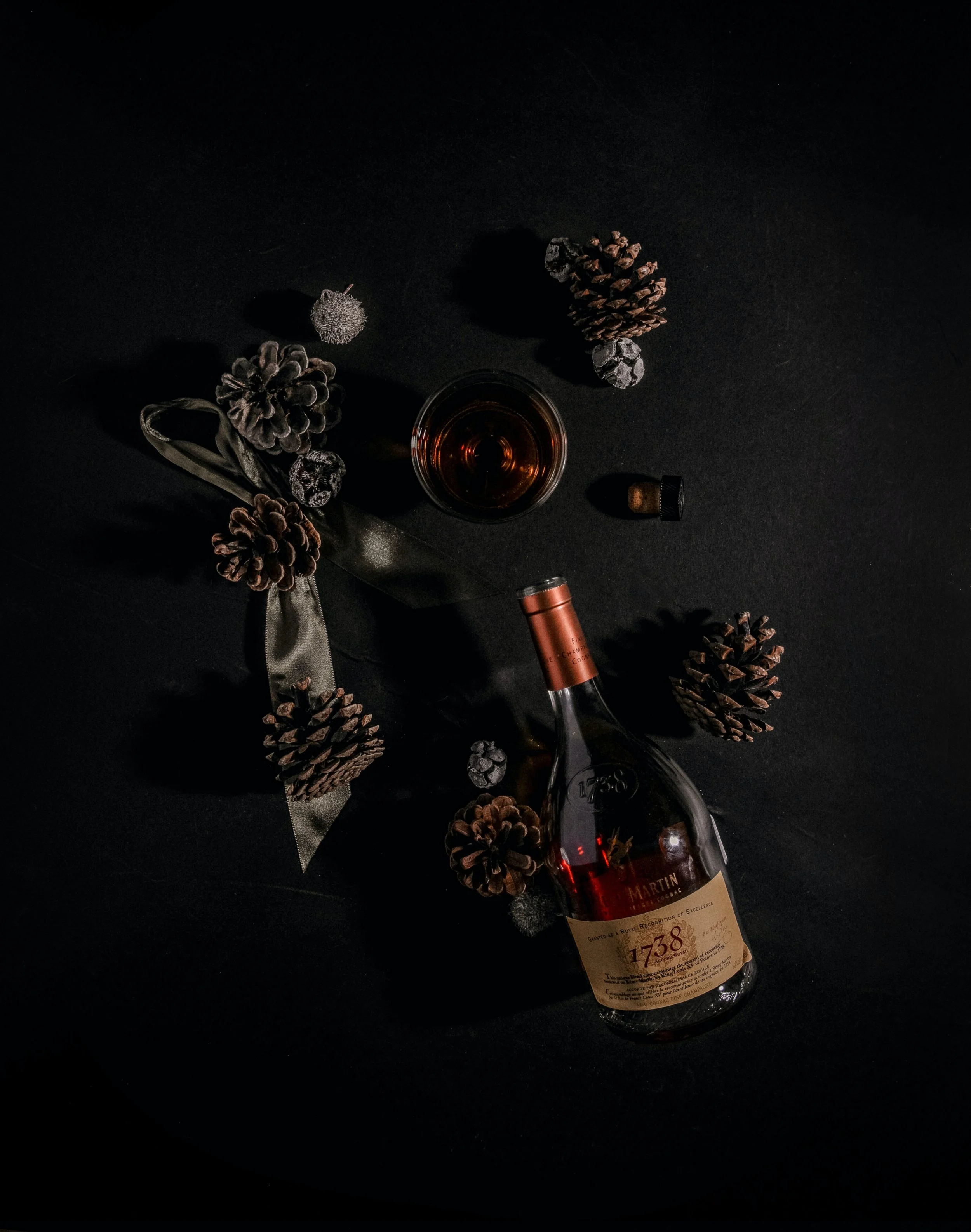A bottle of Martin 1738 wine, a glass of dark red wine, pinecones, a ribbon, a cork, and small decorative rocks arranged on a dark surface.