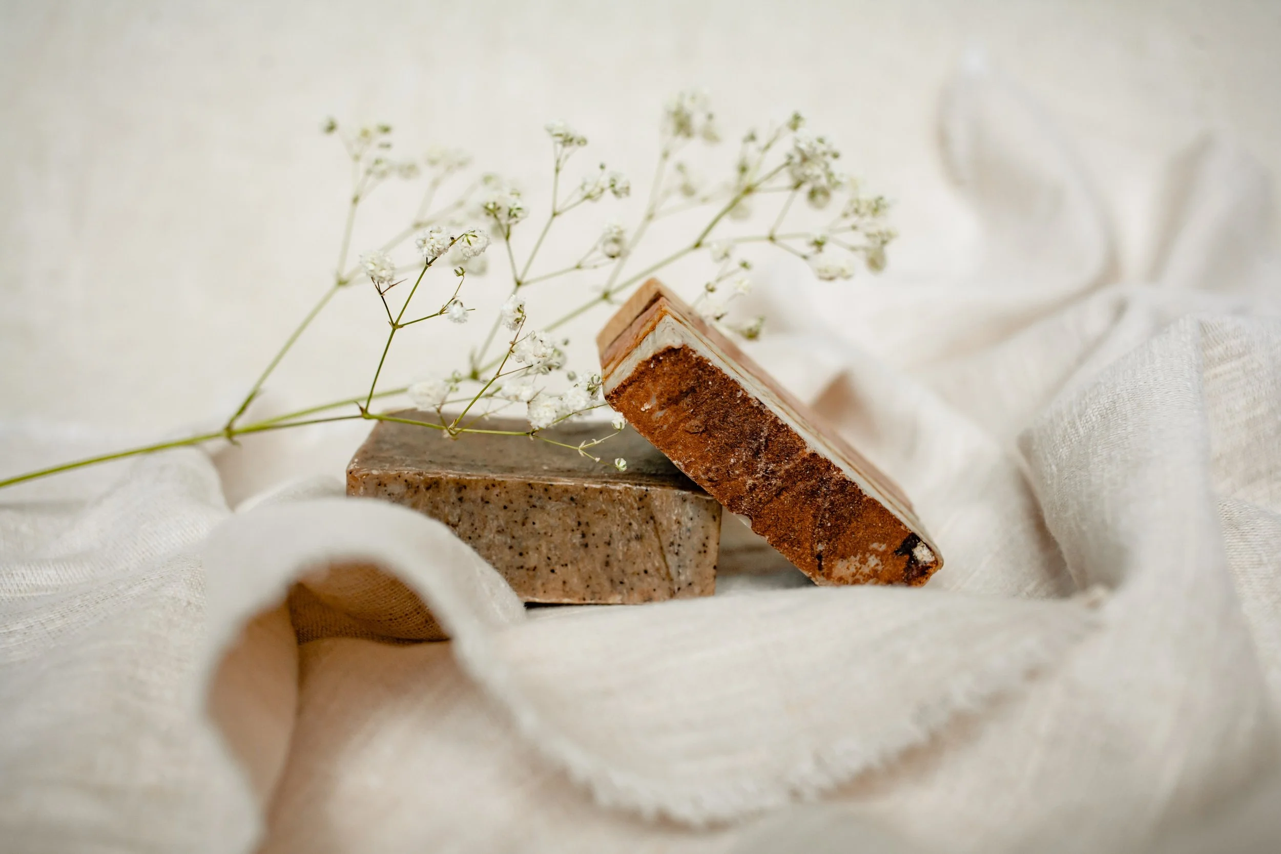 Two handmade soap bars, one beige with speckles and the other darker with a rough texture, are arranged on a soft, cream-colored fabric. A sprig of small white flowers rests on the soaps, creating a natural and rustic aesthetic.