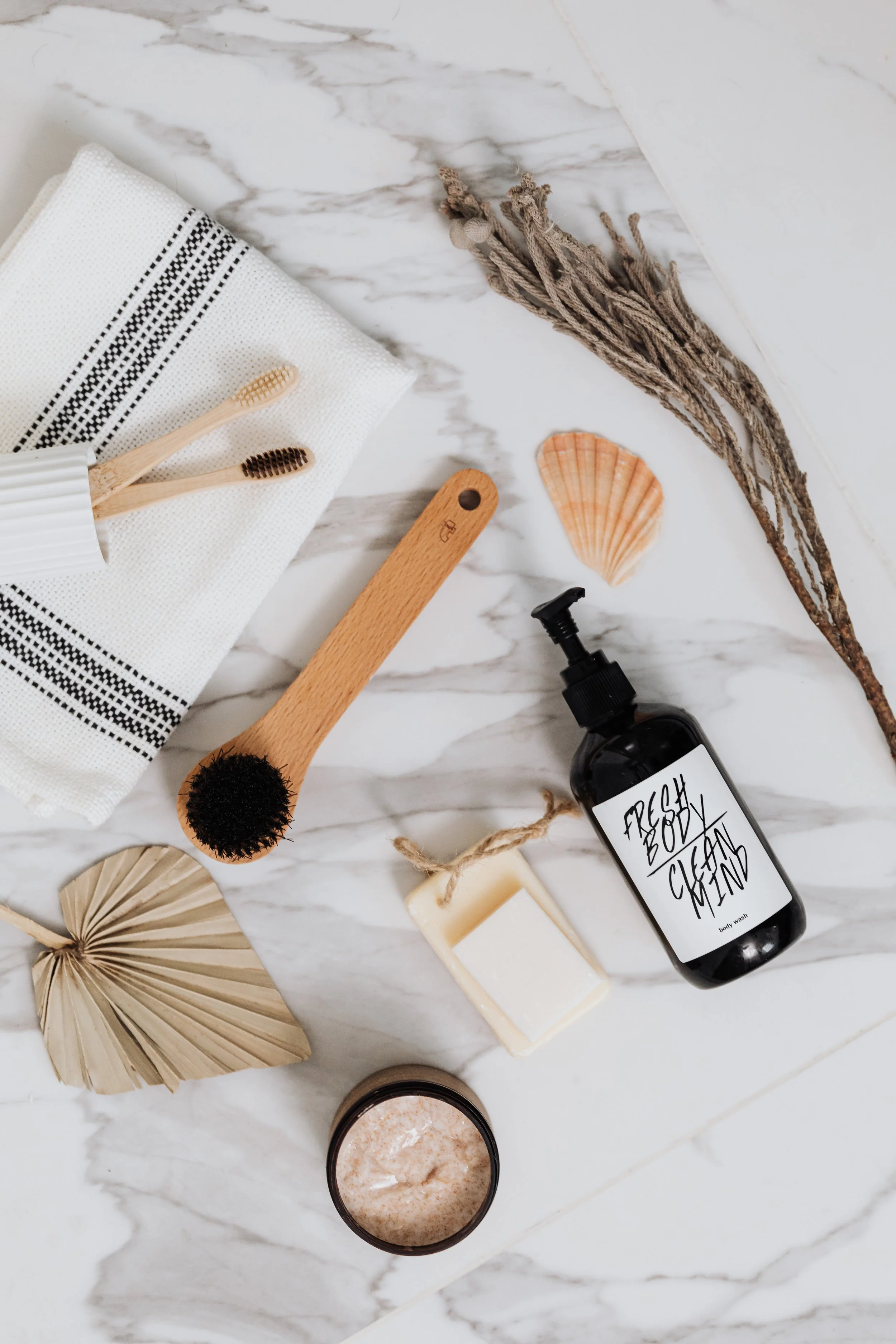 Assorted bathroom and skincare products on a marble countertop including soap, body wash labeled 'fresh box clean', a jar of bath salts, natural body brush, seashells, and dried plants.
