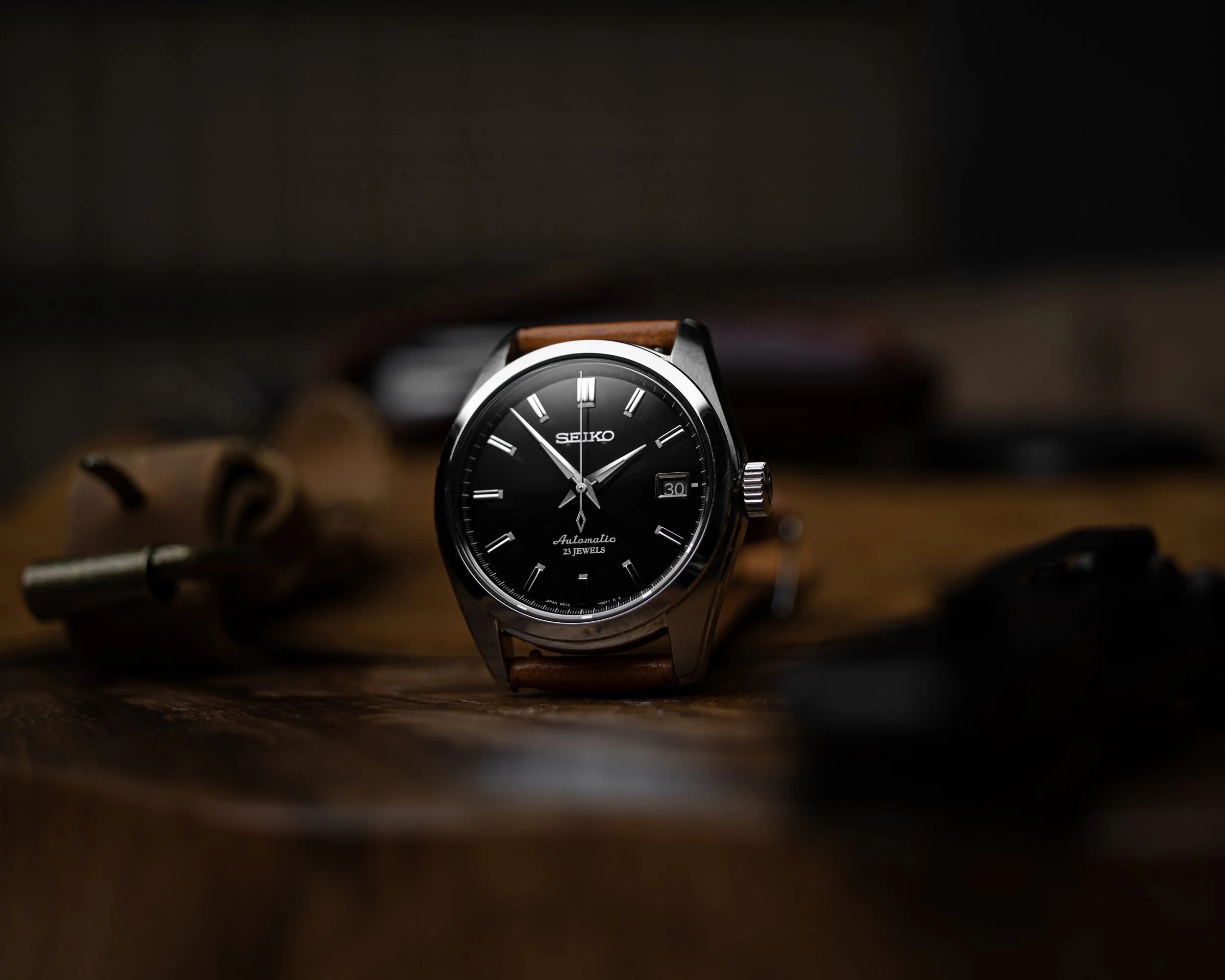 A Seiko automatic wristwatch with a black dial, silver accents, and a brown leather strap, placed on a wooden surface with blurred objects in the background.
