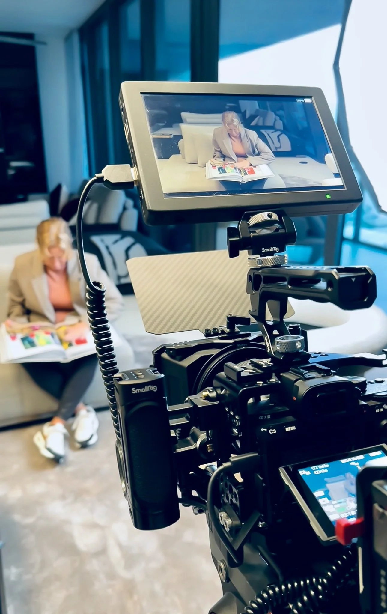 A woman sitting on a couch reading a magazine, being filmed by a professional video camera with a monitor showing the woman in the background.