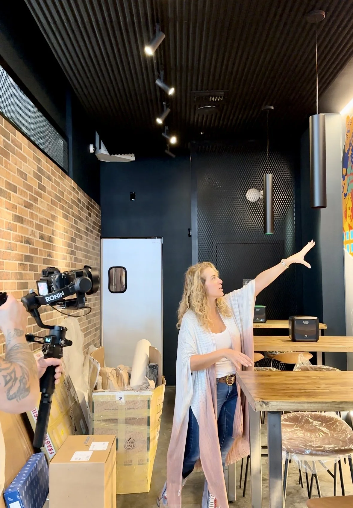 A woman with curly blonde hair standing in a modern indoor space, pointing towards the ceiling while being filmed with a camera operator nearby. The room has a brick wall, black ceiling, and wooden furniture.